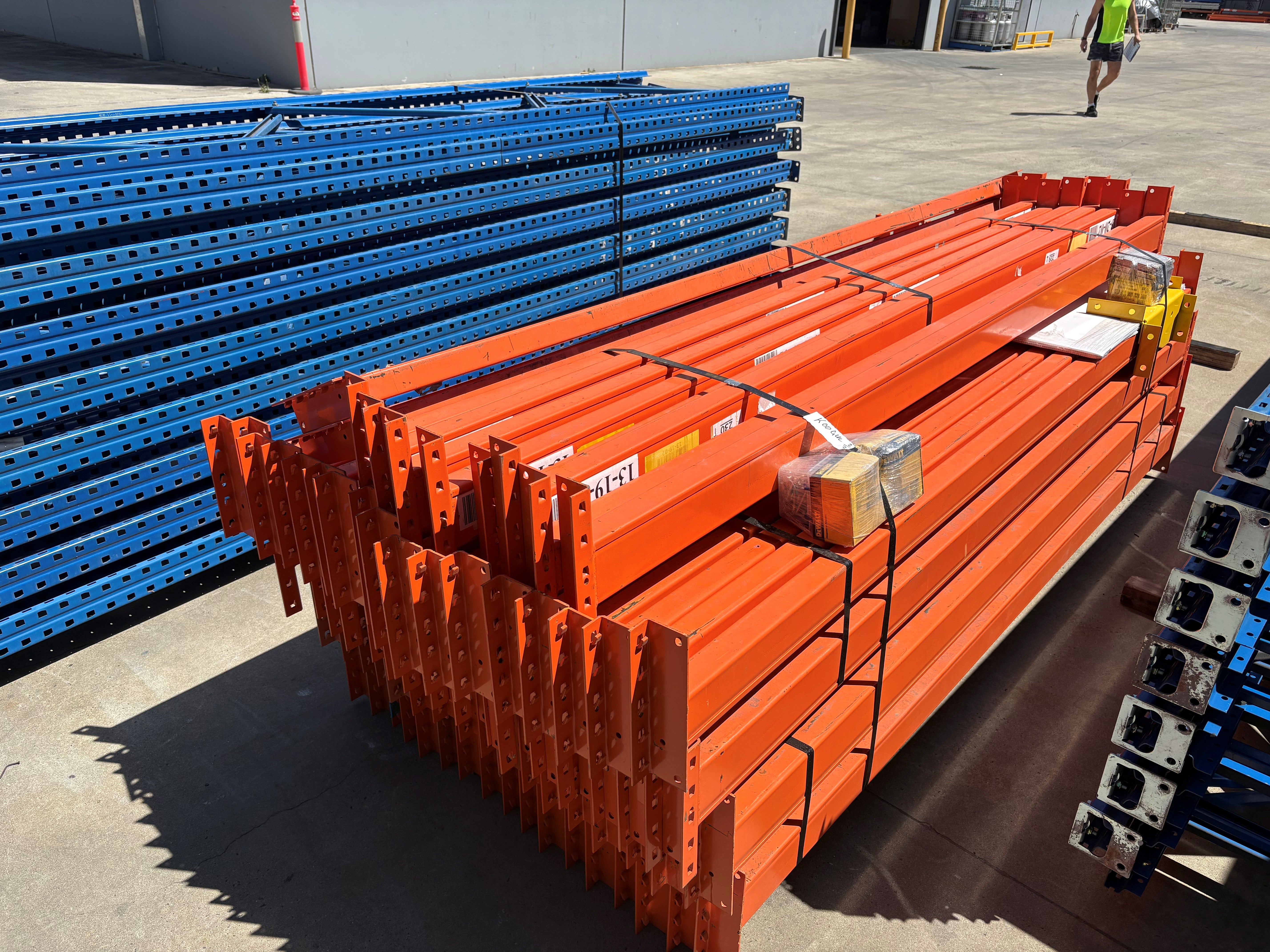 Stacks of orange and blue metal pallets on a concrete surface with a person walking in the background. Schaefer used second hand pallet racking beams and frames 
